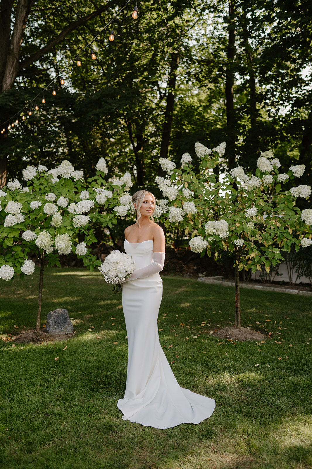 Full-length portrait of the bride holding a white bouquet in a garden with blooming hydrangeas.